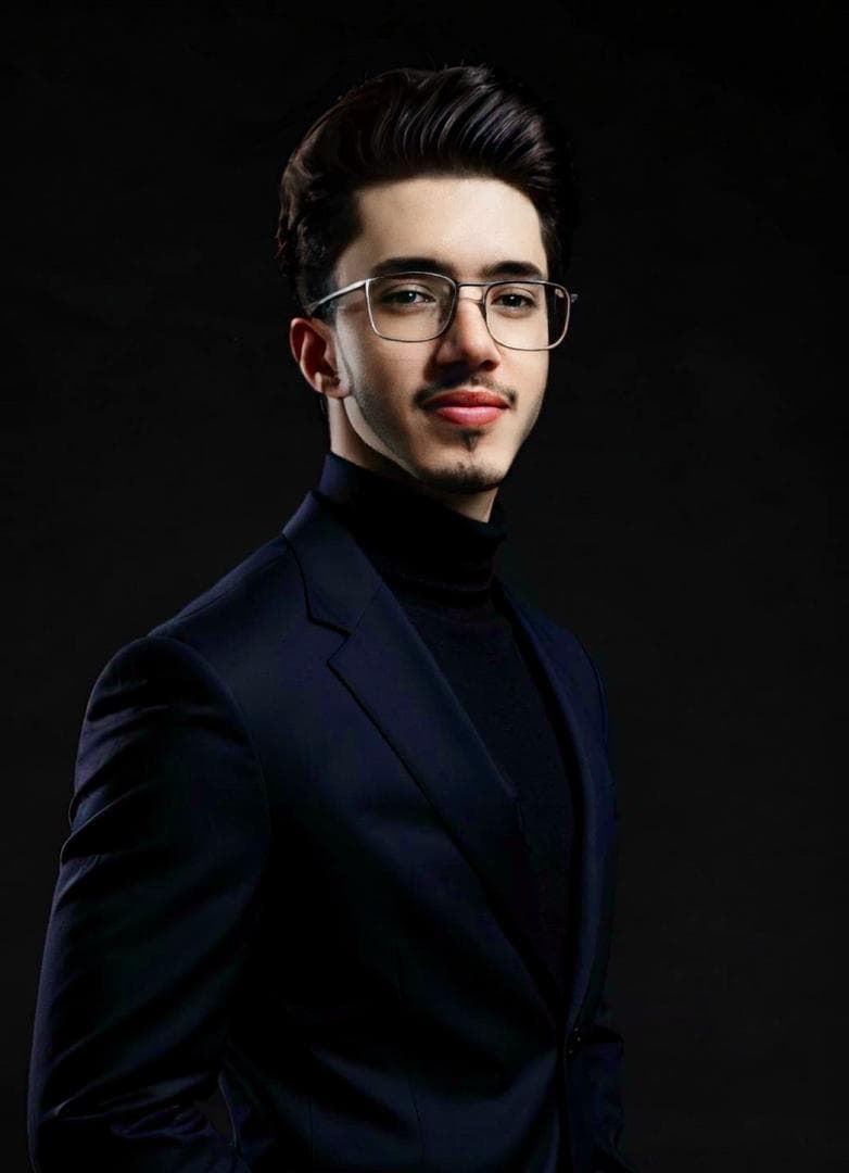 Young man with glasses and mustache wearing a navy suit and black turtleneck against black.